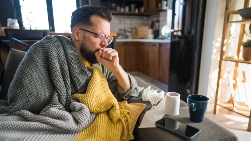 A man sits on a couch bundled up in blankets with tissues and a cup of tea.