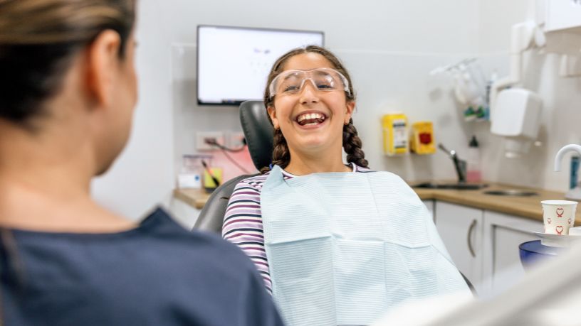 A young girl smiles, showing off her teeth to her dentist as she sits in a chair wearing glasses and a bib.