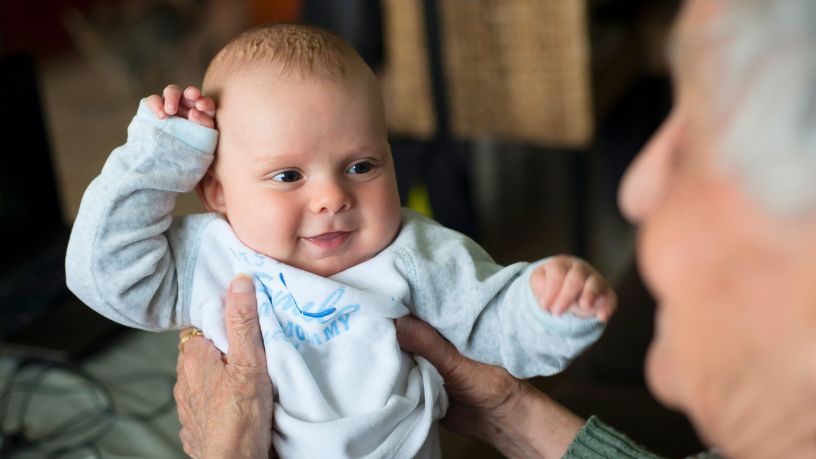 A woman sits in her bathroom holding a small toothbrush in front of her smiling baby.