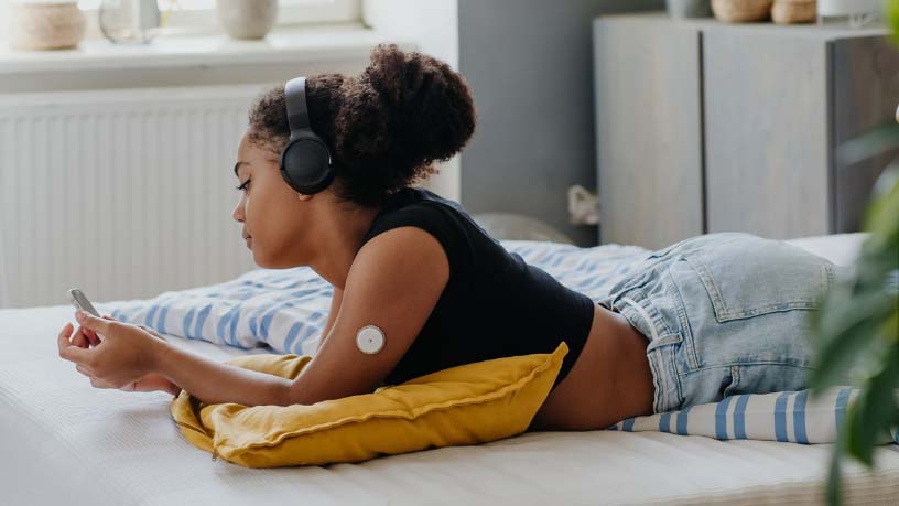 A woman with a continuous glucose monitor on her arm lies on her bed looking at her phone.