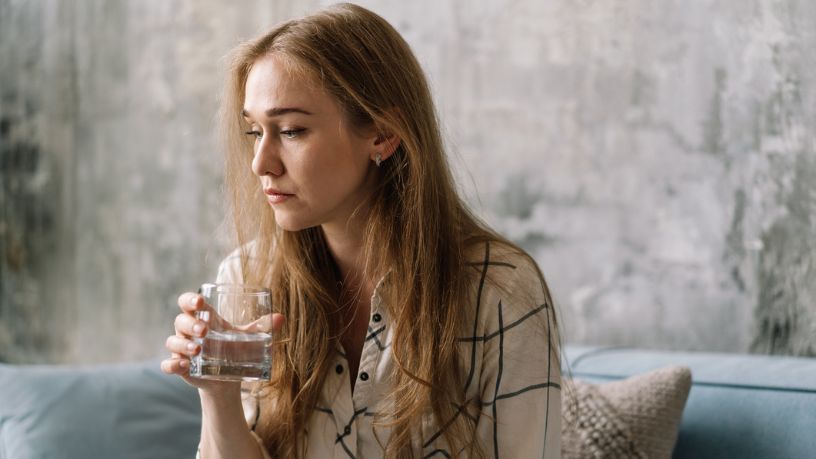 A woman sitting on a couch not looking well/happy whilst drinking water