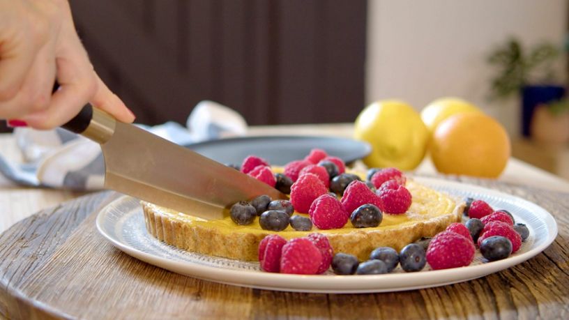 A person slices a lemon cheesecake covered in blueberries and raspberries.
