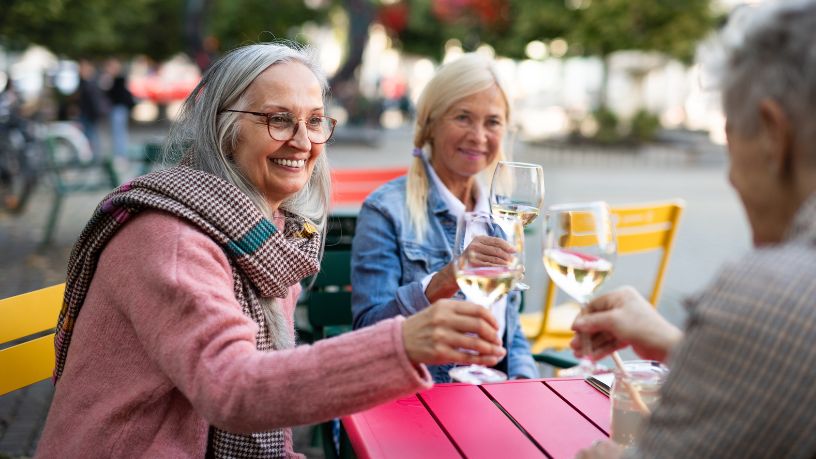 Three older friends toast each other with wine glasses as they sit at an outdoor table.