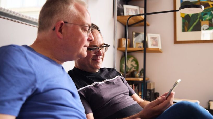 Two men sitting on the couch looking at a mobile phone together.
