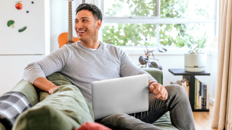 A man sits on a couch with his laptop and smiles at someone out of the shot.