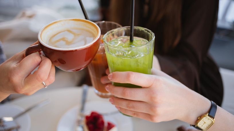 Close-up of three women’s hands clinking glasses of iced matcha latte, espresso tonic and a cappuccino
