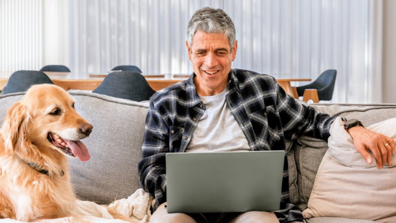 A man sits next to his dog on a couch and works on his laptop.