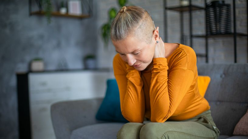 A woman on a couch leans forward and holds her neck.