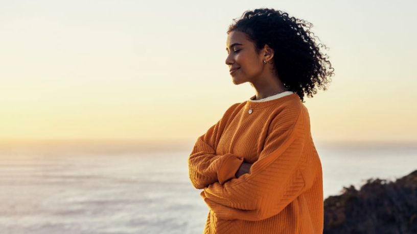 Women stands by the ocean with eyes closed and arms wrapped around her waist.