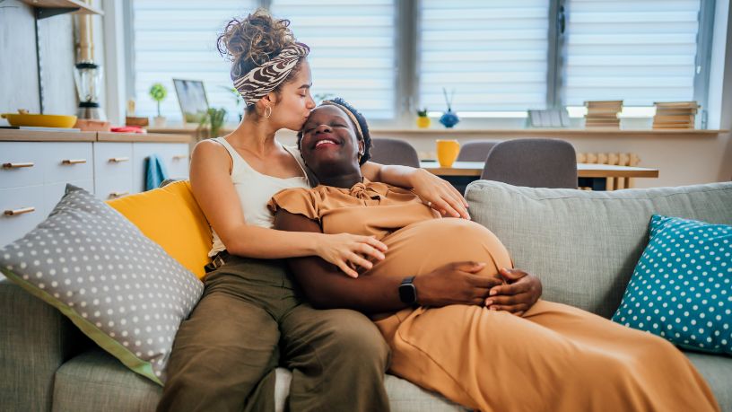 A pregnant woman and her partner laugh and cuddle on their couch.