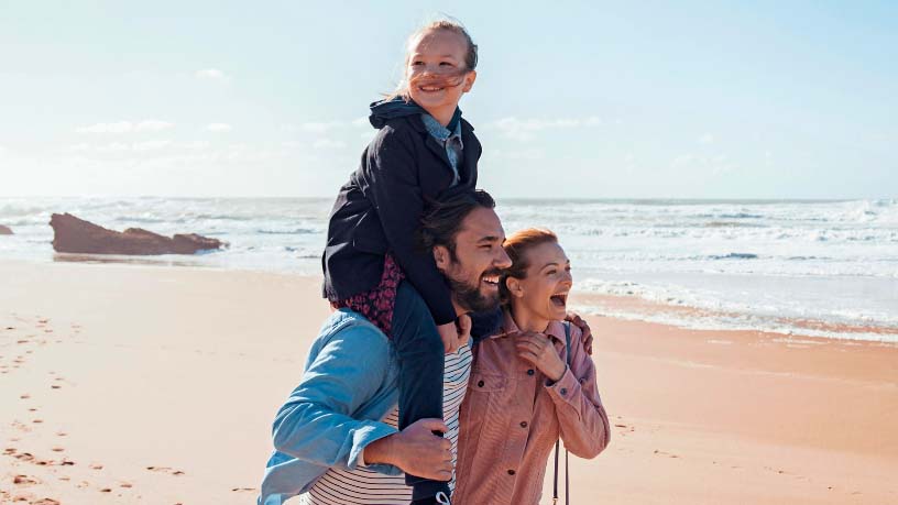 A mum and dad walk along the beach while their daughter sits on her dad’s shoulders.