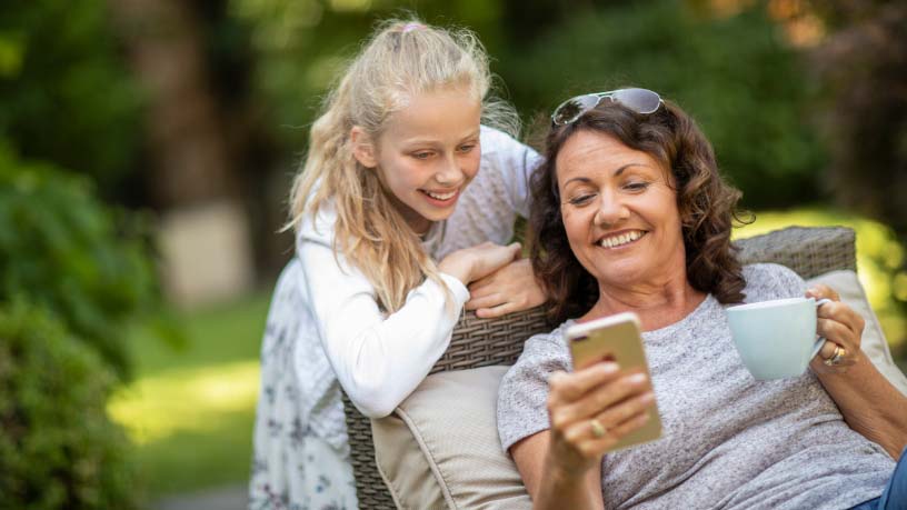 A woman sitting outside grins at her phone with her daughter.