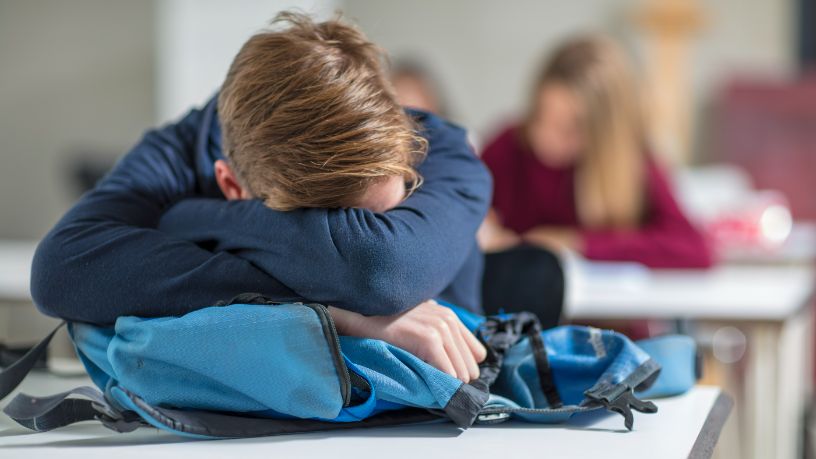 A tired teenager rests on his arms at his classroom desk.