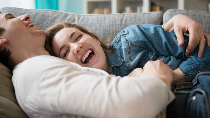 A man and woman cuddle and laugh on their couch.