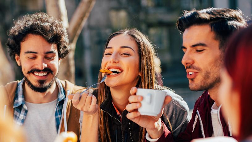Three friends laugh as they eat and drink in a park.