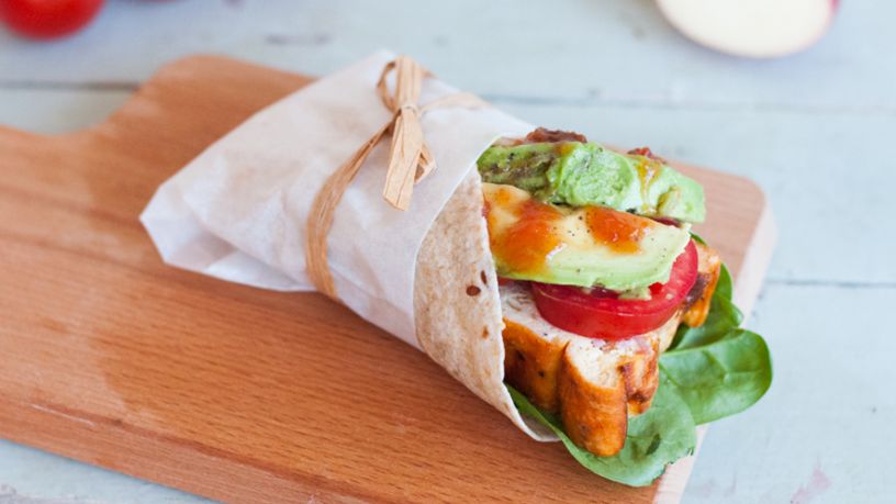 A bacon and egg frittata wrap that’s wrapped in wax paper lays on a serving board.