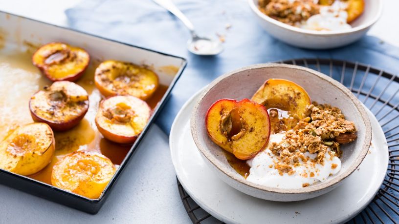Two bowls of spiced stone fruit with yoghurt and granola lay next to a tray of baked nectarines.