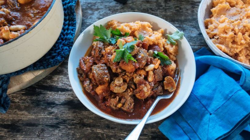 A hearty bowl of beef stew lays on a table next to a larger pot of stew and a dish of sweet potato mash.