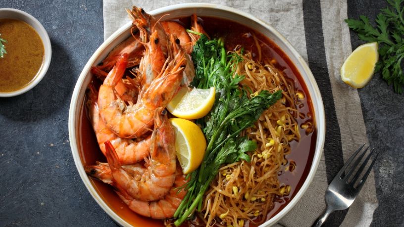 A bowl of prawn laksa sits on a table next to various garnishments.