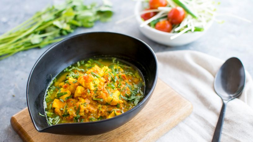 A bowl of fish curry sits on a table next to garnishes and a spoon.