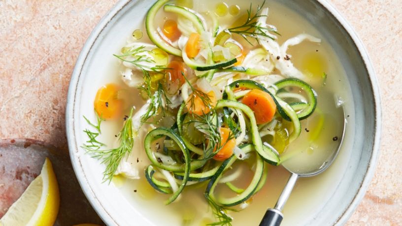 A comforting bowl of chicken soup rests on a dining table with a slice of lemon on the side.