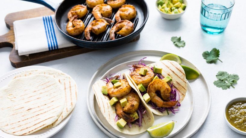 A plate with 2 grilled prawn tacos sits on a dining table alongside a pan with grilled prawns plus various accompaniments.