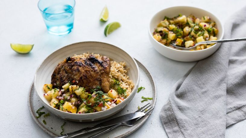 A bowl of jerk chicken thighs on rice sits next to a bowl of pineapple salsa.