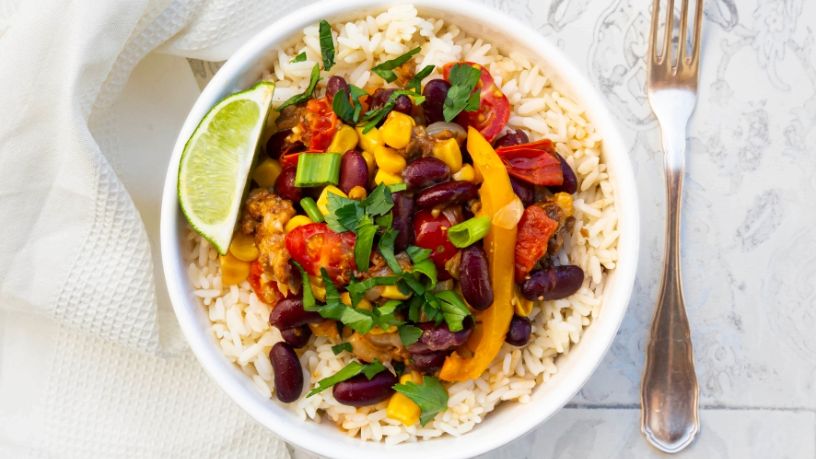 A bowl of rice topped with beans, vegetables and herbs with a wedge of lime on the side