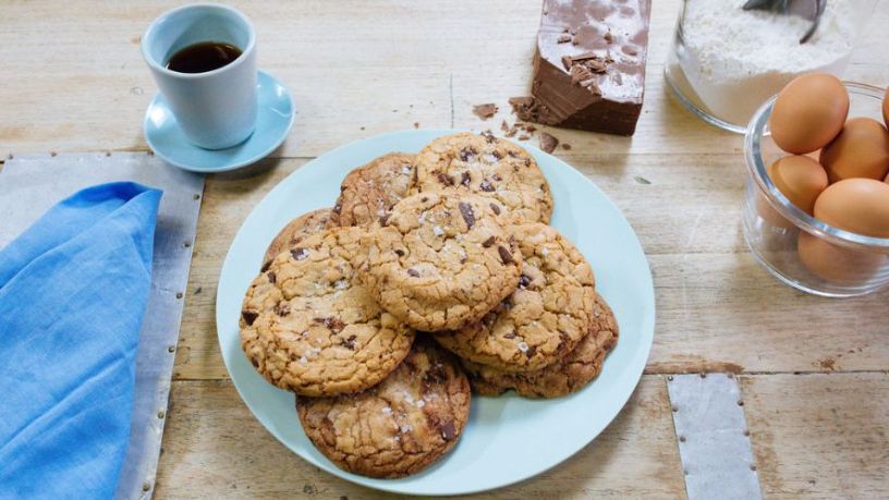 A plate of choc chip cookies lays on a table alongside various cooking ingredients and a cup of black coffee.