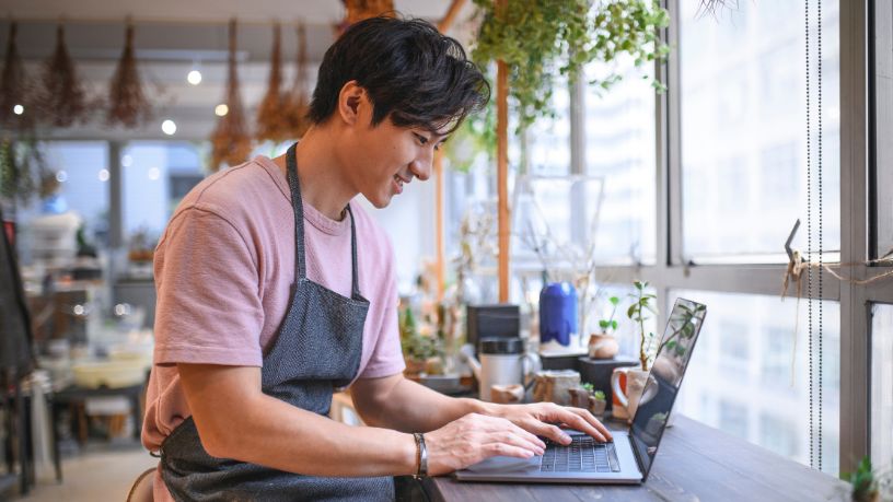 A man wearing an apron sits at a bench in a cafe using his laptop.