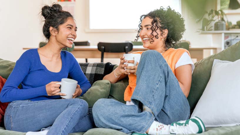 Two women laugh while drinking coffee on a couch.