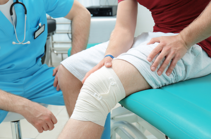 A patient with a bandage over their knee sitting on a doctors bed next to a doctor wearing scrubs