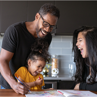 A man with glasses, a child in a yellow shirt, and a woman laughing together in the kitchen while looking at drawings on the table.