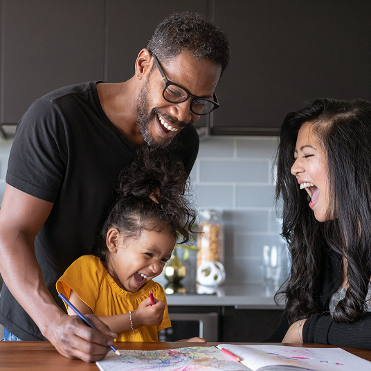 A man with glasses, a child in a yellow shirt, and a woman laughing together in the kitchen while looking at drawings on the table.